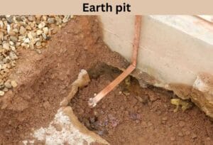 An earth pit with exposed soil and a vertical copper grounding rod connected to a copper strap. Surrounding area has gravel and concrete.